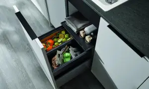 Wysuwana szuflada z podwójnym koszem na śmieci, w której widać owoce, warzywa i papier. Obok leżą butelki i ręcznik.