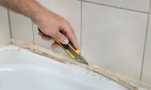 Ręka z nożem odcina stary silikon, pokazując, jak usunąć stary silikon z wanny.