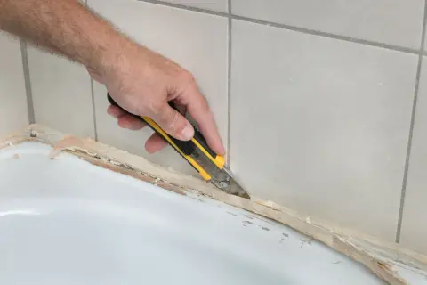 Ręka z nożem odcina stary silikon, pokazując, jak usunąć stary silikon z wanny.