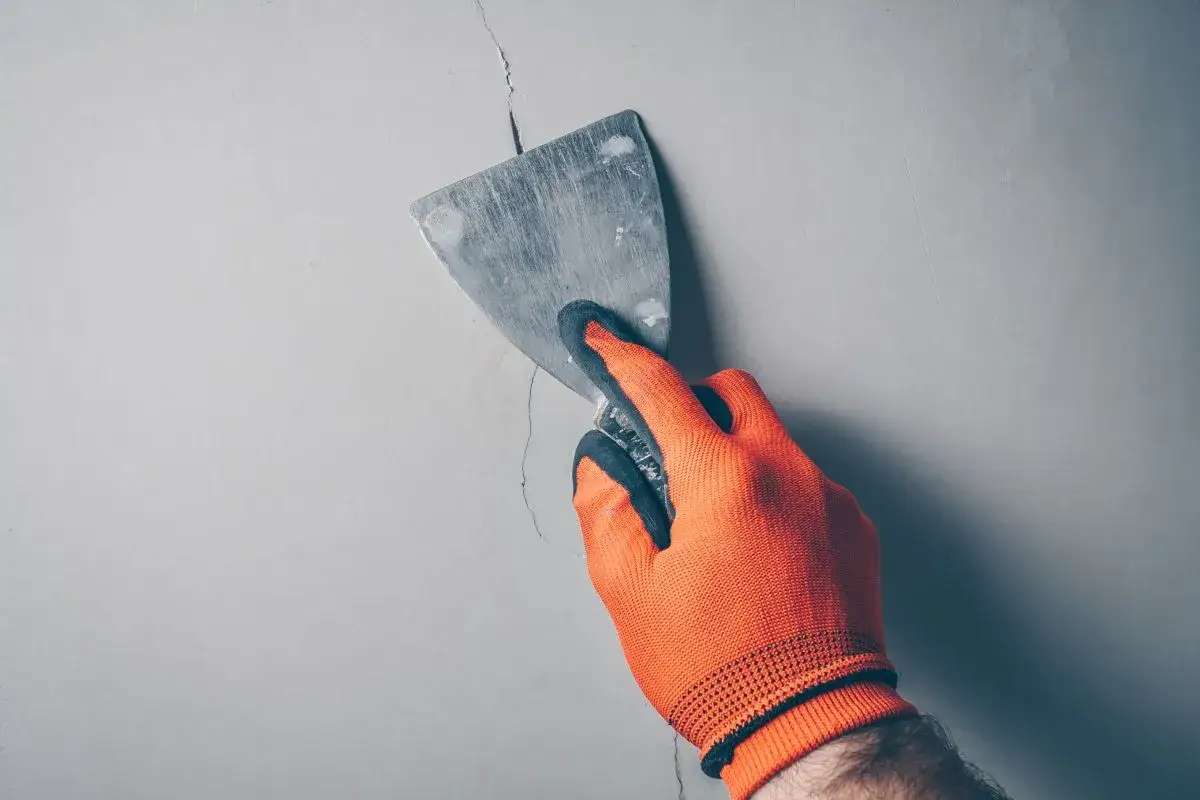 Ręka w pomarańczowej rękawicy zacierająca pęknięcie na ścianie za pomocą szpachli. Naprawa pęknięć ścian.